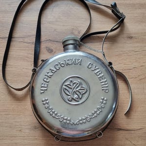 Op de afbeelding: Een ronde, zilverkleurige metalen veldfles met een zwarte lederen schouderriem. De voorzijde is voorzien van een centraal bloemembleem in reliëf, een decoratieve rankenrand onderaan en de cyrillische tekst "ЧЕРКАСЬКИЙ СУВЕНІР" bovenaan.