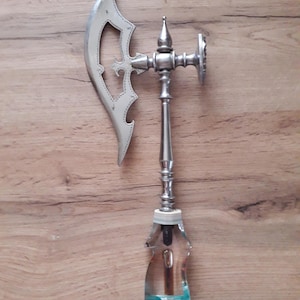 May include: A decorative bottle stopper with a silver axe-shaped top and a clear, faceted glass base. The axe head has a detailed design with a cross motif. The stopper is designed to fit into a bottle.
