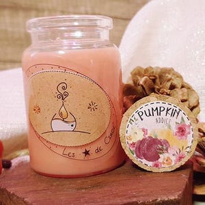 Peut inclure: Une bougie en verre avec une cire rose et orange. Le bocal a une étiquette avec une illustration dessinée à la main d'une flamme et le texte "Les Etoiles de Cherie". Le bocal est posé sur une surface en bois avec une petite citrouille et des pommes de pin.