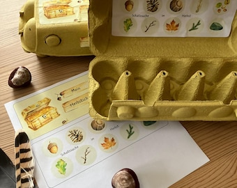 May include: A yellow egg carton with a treasure chest illustration and the words "Mein Herbstschatz". The carton is open, revealing egg holders and a sheet with nature-themed stickers, including acorns and leaves. A feather and a chestnut are also present.
