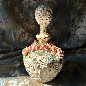 May include: A white porcelain perfume bottle with a floral design. The bottle is decorated with pink and blue flowers and gold accents. The bottle has a decorative stopper with a floral design.