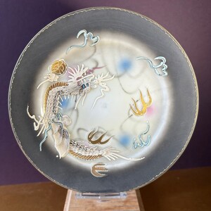 May include: A decorative plate with a gold dragon design on a black background. The dragon is surrounded by clouds and other decorative elements. The plate is on a wooden stand.