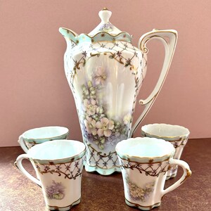 May include: A set of delicate porcelain teacups and a teapot with a floral design. The teapot has a light blue and gold trim with a floral pattern. The teacups have a similar design with a light blue and gold trim. The cups and teapot are on a brown surface.