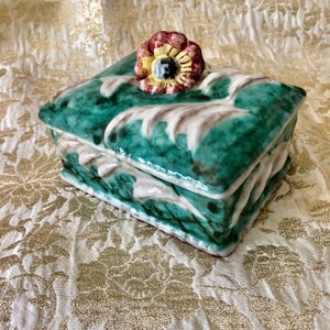 May include: A green ceramic trinket box with a white floral design and a red, yellow, and black flower on the lid.