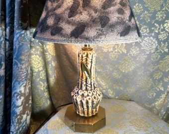 May include: A table lamp with a bone-shaped ceramic base and a leopard print lampshade. The base is decorated with green leaves and the lampshade is a light brown color with black spots.