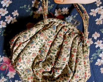 May include: A small, round, cream-coloured purse with a floral pattern. The purse has a drawstring closure and a long strap.