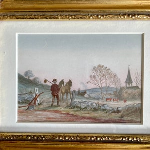 May include: A framed watercolor painting depicting a rural scene with a man leading two horses down a path towards a church with a bell tower. The sky is a pale blue with a crescent moon visible. The painting is set in a gold frame.