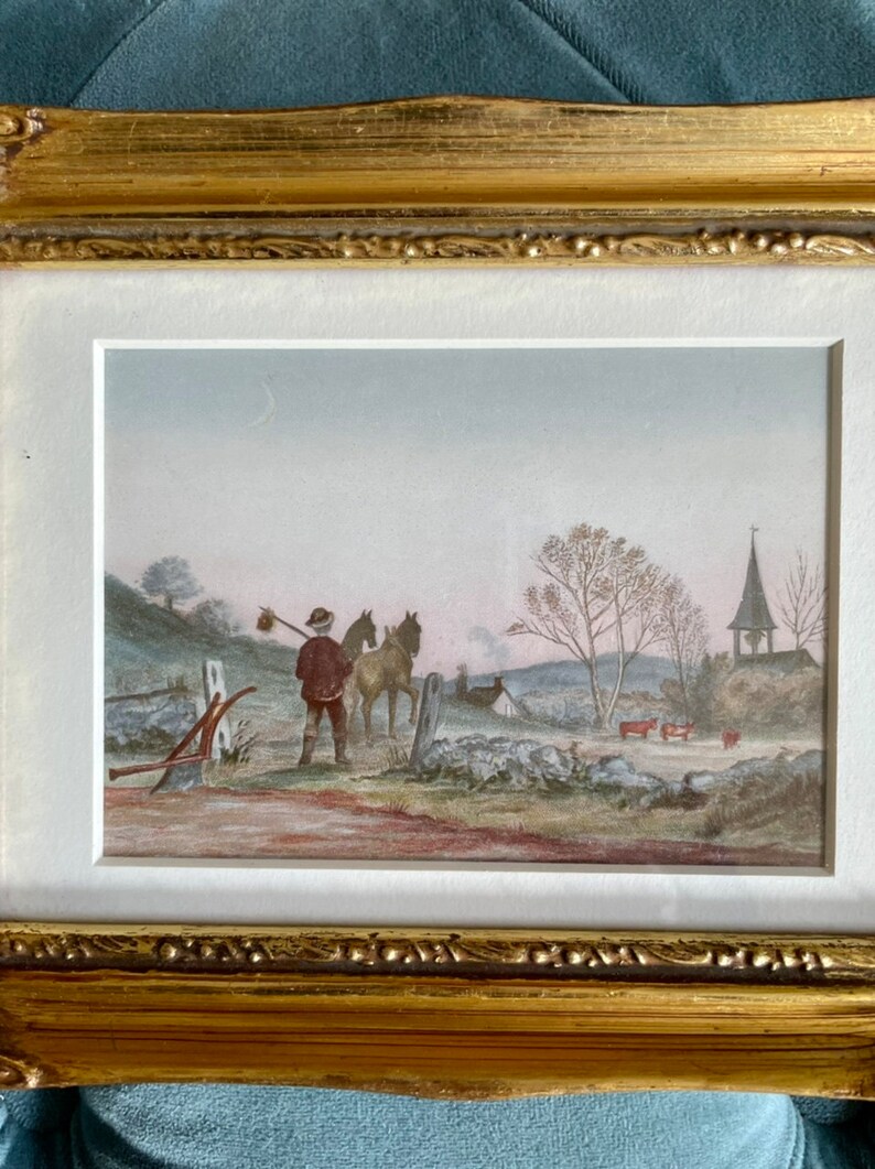 May include: A framed watercolor painting depicting a rural scene with a man leading two horses down a path towards a church with a bell tower. The sky is a pale blue with a crescent moon visible. The painting is set in a gold frame.