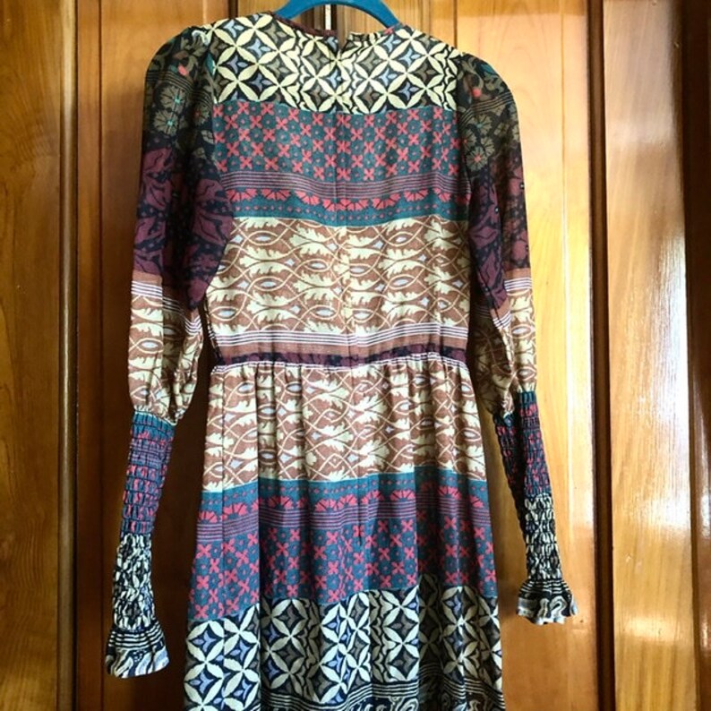 May include: A long-sleeved dress with a colorful geometric pattern. The dress has a fitted bodice and a flowing skirt. The fabric is sheer and lightweight.