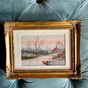 May include: A framed watercolor painting depicting a rural landscape with a river, trees, and a small cottage. The painting is set in a soft, muted palette of browns, greens, and blues, with a hint of pink in the sky. The painting is framed in a gold-colored frame.