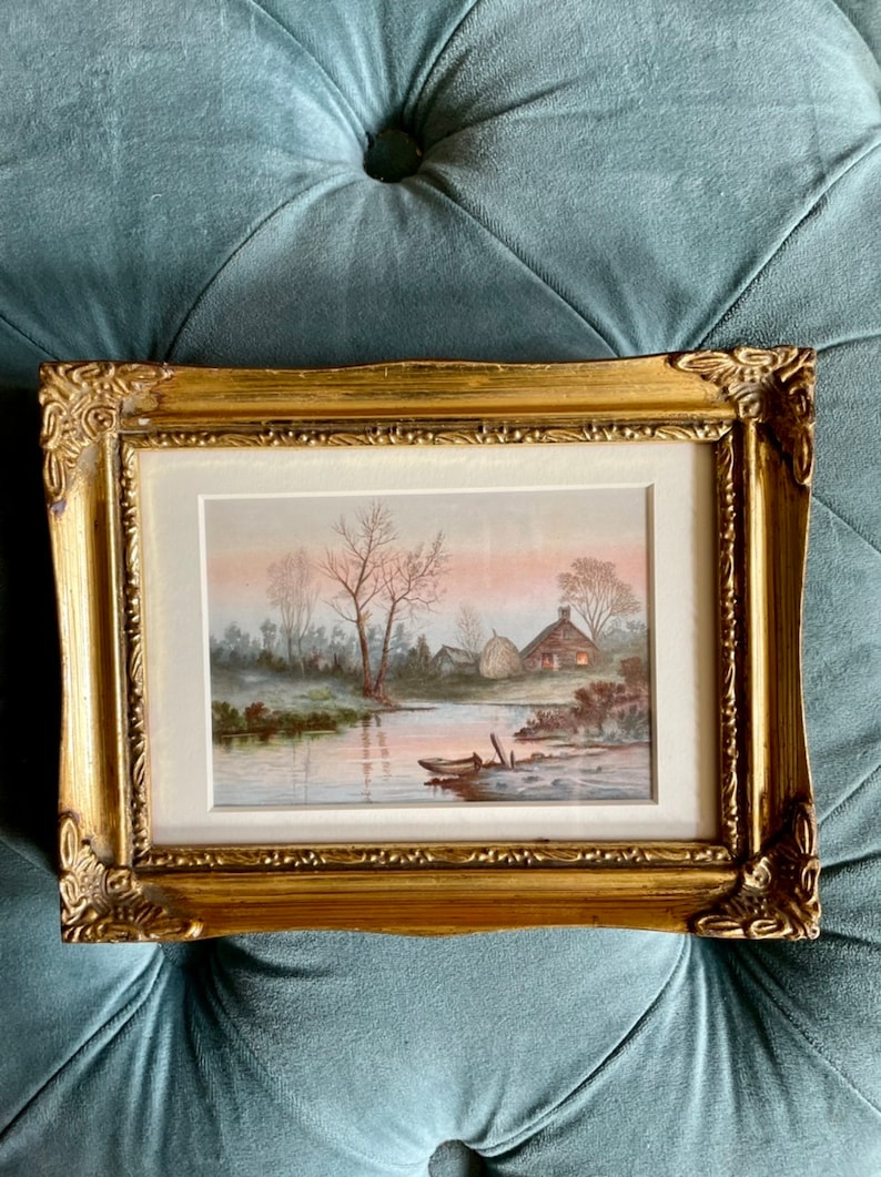May include: A framed watercolor painting depicting a rural landscape with a river, trees, and a small cottage. The painting is set in a soft, muted palette of browns, greens, and blues, with a hint of pink in the sky. The painting is framed in a gold-colored frame.