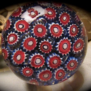 May include: A glass paperweight with a swirling pattern of red, white, and blue. The paperweight is round and has a smooth, glossy finish.
