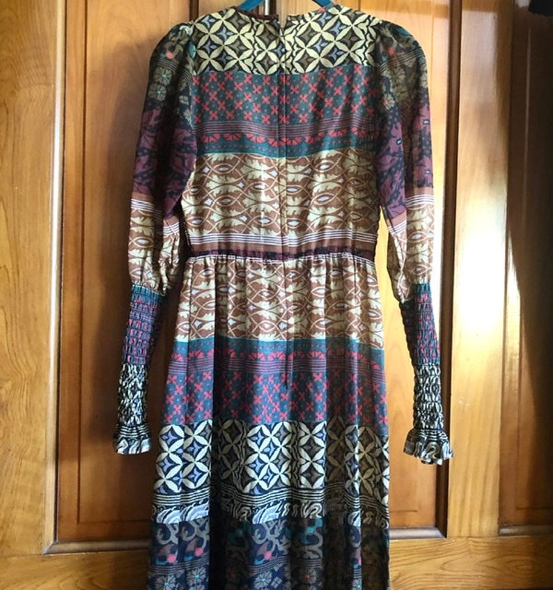 May include: A long-sleeved dress with a colorful, geometric pattern. The dress has a fitted bodice and a flowing skirt. The fabric is sheer and lightweight.
