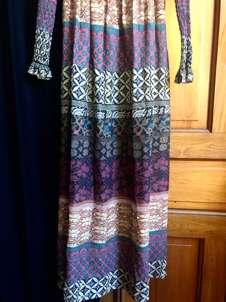 May include: A long, flowing maxi dress with a colorful, geometric pattern. The dress has a fitted bodice and a flowing skirt with a variety of colors, including brown, blue, red, and white.