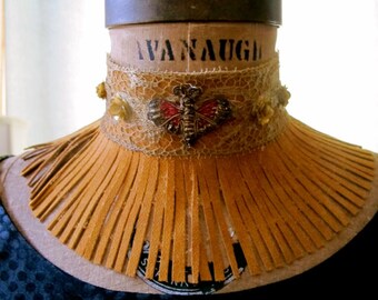 May include: A tan leather collar with fringe detail. The collar features a lace overlay with a red and silver butterfly brooch and small yellow floral accents. The collar is displayed on a vintage dress form.