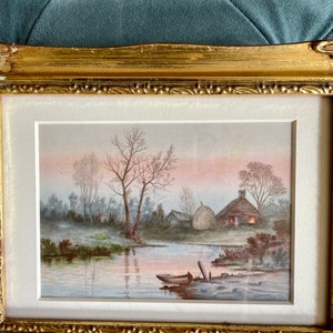May include: A framed watercolor painting depicting a rural scene with a small cottage, a hay bale, and a boat on a river. The sky is a soft pink and the trees are bare.