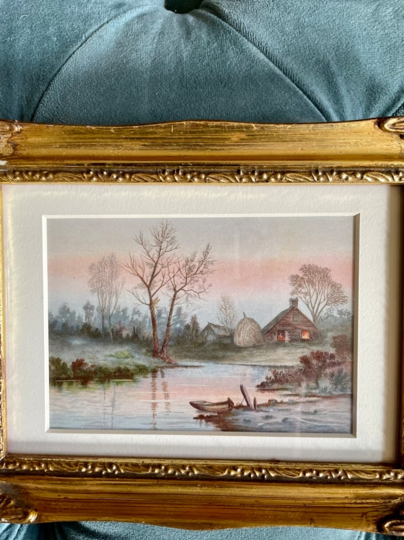 May include: A framed watercolor painting depicting a rural scene with a small cottage, a hay bale, and a boat on a river. The sky is a soft pink and the trees are bare.