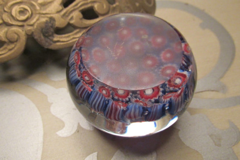 May include: A round glass paperweight with a swirling pattern of blue, red, and white. The paperweight has a clear glass base and a cloudy, white center.
