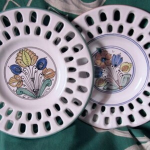 May include: Two white ceramic plates with a decorative floral design. The plates have a scalloped edge and a cut-out pattern around the rim.