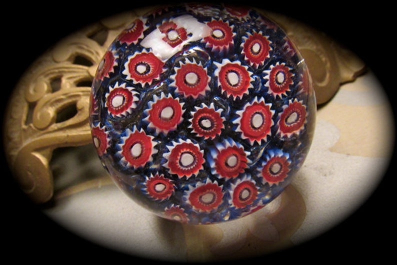 May include: A glass paperweight with a red, white, and blue pattern. The pattern is made up of small, circular designs.
