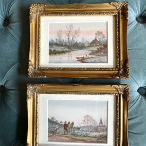 May include: Two framed watercolor paintings with gold frames. The top painting depicts a river scene with a small house and trees. The bottom painting depicts a man on horseback riding through a rural landscape.