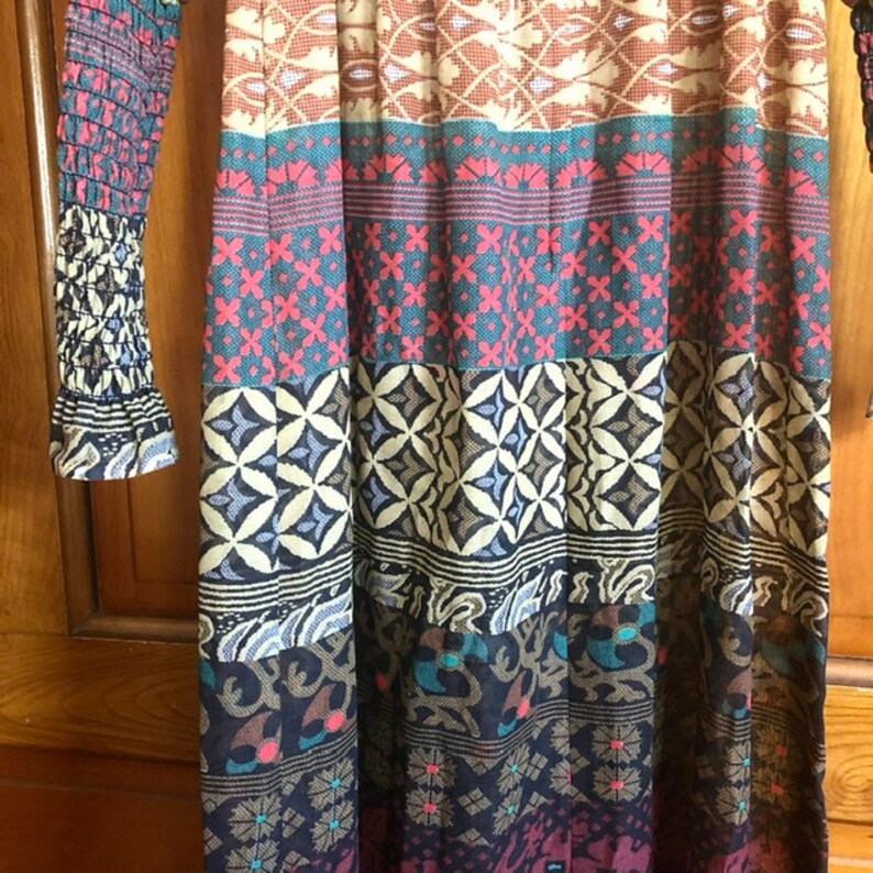 May include: A long, flowing dress with a colorful, patterned design. The dress has a fitted bodice and a flowing skirt with multiple layers of fabric. The pattern features a variety of geometric shapes and floral motifs in shades of brown, blue, red, and white.