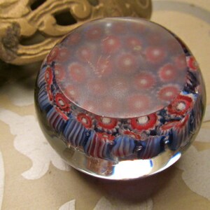 May include: A clear glass paperweight with a swirling pattern of red, white, and blue. The paperweight has a clear, round shape and is filled with small, colorful bubbles.