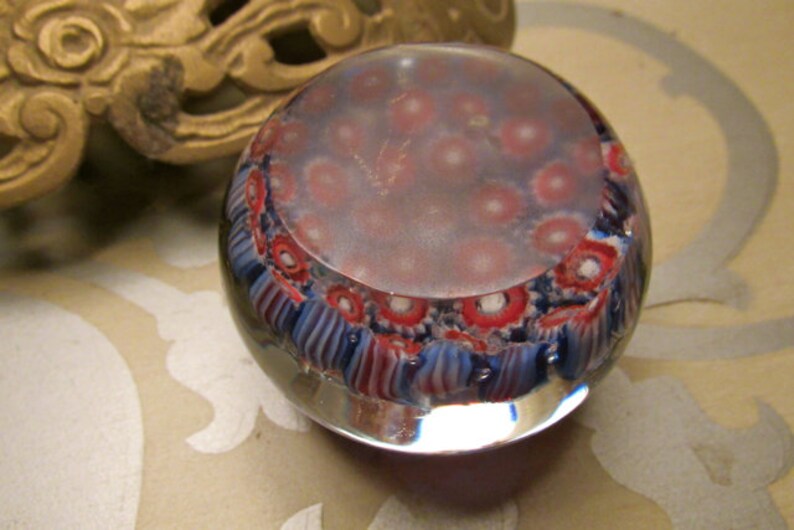 May include: A clear glass paperweight with a swirling pattern of red, white, and blue. The paperweight has a clear, round shape and is filled with small, colorful bubbles.