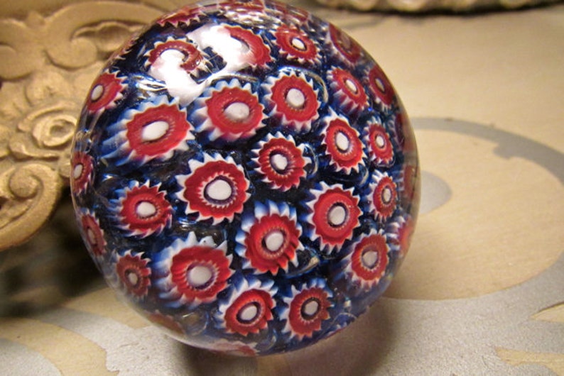 May include: A glass paperweight with a swirling pattern of red, white, and blue. The paperweight is round and has a smooth, shiny surface.