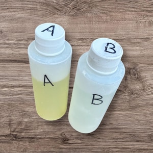 May include: Two clear plastic bottles with white caps labeled "A" and "B". Bottle "A" contains a yellow liquid, while bottle "B" holds a clear liquid. The bottles are on a wooden surface.