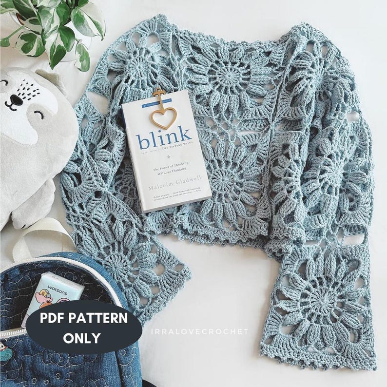May include: A light blue crochet top with floral patterns and bell sleeves. A book titled "blink" is placed on the top. A gold heart-shaped bookmark is attached to the book. A denim backpack with the text "PDF PATTERN ONLY" is in the image.