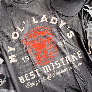 May include: A dark gray t-shirt with the text "MY OL' LADY'S BEST MISTAKE" and a black cap with a white logo. Tools like a wrench, hammer, and pliers are also visible, along with a can.