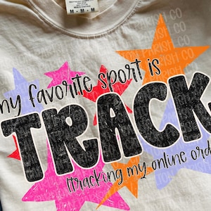 May include: A cream-colored short-sleeve t-shirt with a graphic print featuring orange, pink, and purple stars. The text reads: my favorite sport is TRACK (tracking my online orders). The shirt has a Comfort Colors brand label at the neck.