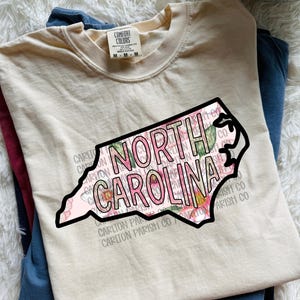 May include: A cream-colored t-shirt with a graphic of the North Carolina state outline, filled with pink floral patterns and the words "NORTH CAROLINA" in a bold font. The shirt is folded with other shirts.