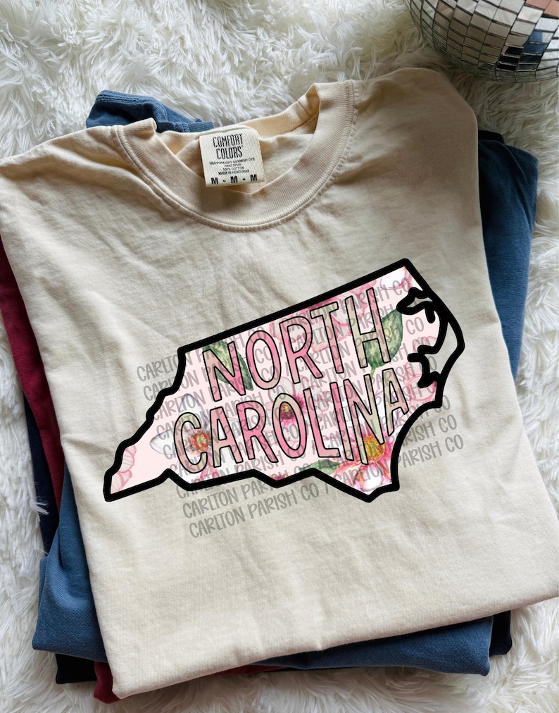 May include: A cream-colored t-shirt with a graphic of the North Carolina state outline, filled with pink floral patterns and the words "NORTH CAROLINA" in a bold font. The shirt is folded with other shirts.
