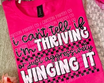 May include: A bright pink t-shirt featuring the black and white text "I can't tell if I'm THRIVING or just aggressively WINGING IT" layered over a black and white checkered pattern. The shirt has a crew neckline and a black brand label at the collar.