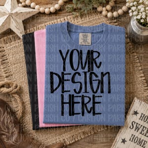 May include: Three t-shirts in black, pink, and blue, folded and stacked on a burlap surface. The blue shirt has the text "YOUR DESIGN HERE" in large black letters. A wooden sign with "HOME SWEET HOME" is visible.