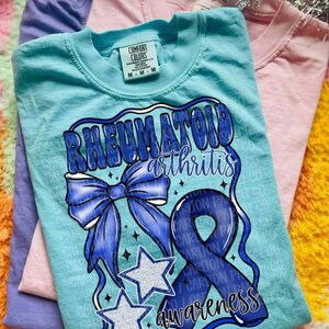 May include: A light blue short-sleeve t-shirt featuring a graphic print with the text "RHEUMATOID arthritis awareness". The design includes a large blue bow, a blue awareness ribbon, and two white stars with blue outlines, all set against a light blue fabric.