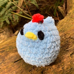May include: A blue crocheted bird with a red pom-pom on its head. The bird has black eyes and a yellow beak. It is sitting on a brown log.