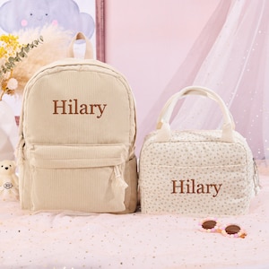 K&ouml;nnte beinhalten: Ein beiges Cord-Rucksack- und Lunchpaket-Set. Der Rucksack hat eine Vordertasche und den Namen "Hilary" in Braun gestickt. Die Lunchtasche hat ein Blumenmuster und den Namen "Hilary" in Braun gestickt.