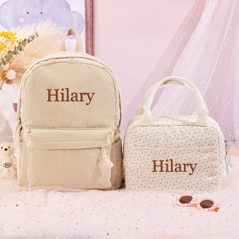 K&ouml;nnte beinhalten: Ein beiges Cord-Rucksack- und Lunchpaket-Set. Der Rucksack hat eine Vordertasche und den Namen "Hilary" in Braun gestickt. Die Lunchtasche hat ein Blumenmuster und den Namen "Hilary" in Braun gestickt.