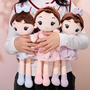 May include: Three plush dolls with brown hair, wearing pink, purple, and blue dresses. The dolls have large eyes and smiling faces. They are being held by a person.