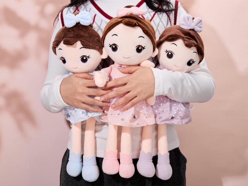May include: Three plush dolls with brown hair, wearing pink, purple, and blue dresses. The dolls have large eyes and smiling faces. They are being held by a person.