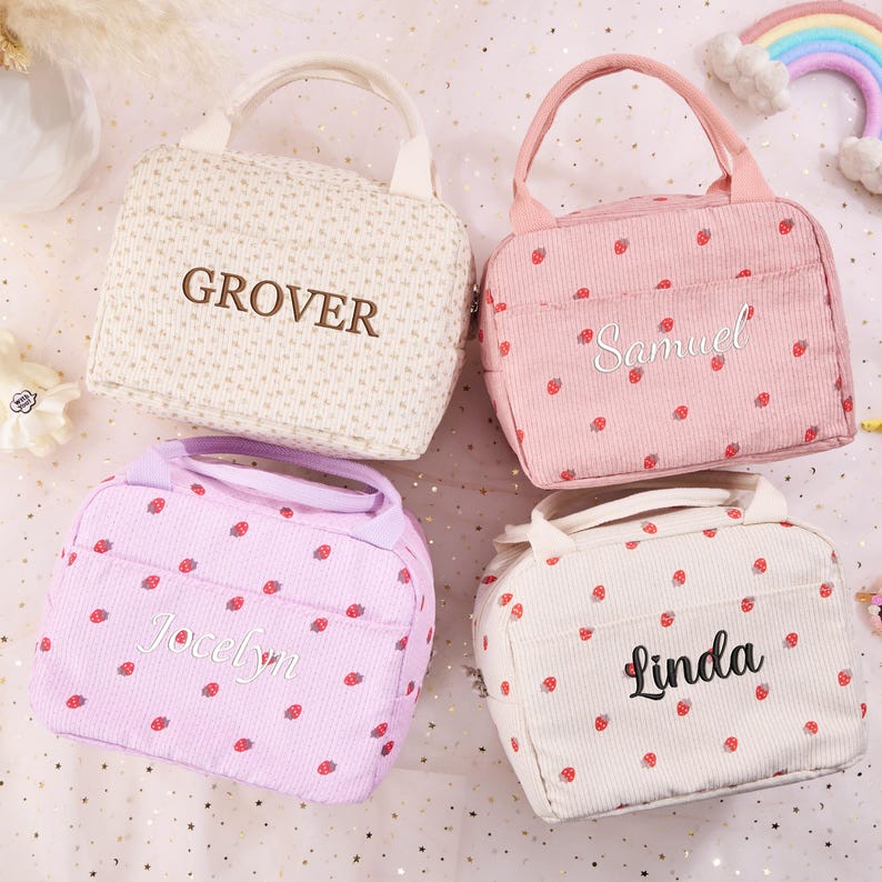 K&ouml;nnte beinhalten: Vier Lunchpakete in Creme, Rosa und Lavendel, jeweils mit Griff und Vordertasche. Die cremefarbene Tasche hat den Namen "GROVER" gestickt. Die rosa Tasche hat den Namen "Samuel" gestickt. Die lavendelfarbene Tasche hat den Namen "Jocelyn" gestickt. Die cremefarbene Tasche hat den Namen "Linda" gestickt. Jede Tasche ist mit kleinen roten Erdbeeren verziert.