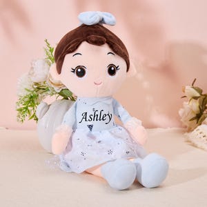 May include: A plush doll with brown hair, a blue bow, and a blue dress with white lace trim. The doll has a smiling face with brown eyes and is wearing a blue dress with the name "Ashley" embroidered on the front.