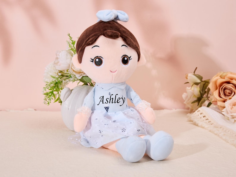 May include: A plush doll with brown hair, a blue bow, and a blue dress with white lace trim. The doll has a smiling face with brown eyes and is wearing a blue dress with the name "Ashley" embroidered on the front.