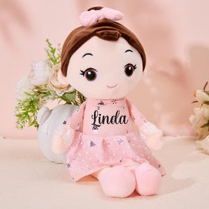 May include: A pink plush doll with brown hair and a pink bow. The doll is wearing a pink dress with white polka dots and the name "Linda" is embroidered on the front.