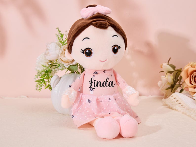 May include: A pink plush doll with brown hair and a pink bow. The doll is wearing a pink dress with white polka dots and the name "Linda" is embroidered on the front.