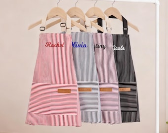Puede incluir: Cuatro delantales personalizados colgados de perchas de madera. Los delantales son a rayas en rojo, gris, rosa y negro. Cada delantal tiene un nombre bordado en la parte delantera: Rachel, Vivía, Tiny y Nicole. Cada delantal tiene un bolsillo de parche de cuero marrón.