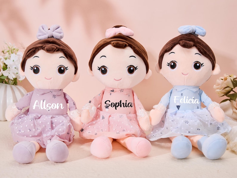 May include: Three plush dolls with brown hair, wearing dresses and bows. The dolls are named Allison, Sophia, and Felicia.